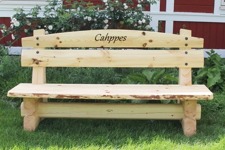Rustik bänk från Nordanlids Rustik med timrat underrede i massivt trä. Bänken har ett vackert utformat ryggstöd som smyckas med dekorativa smidesspik, vilket skapar en gedigen och tidlös känsla av hantverk. Snyggt en din egna infrästa text i ryggstödet.